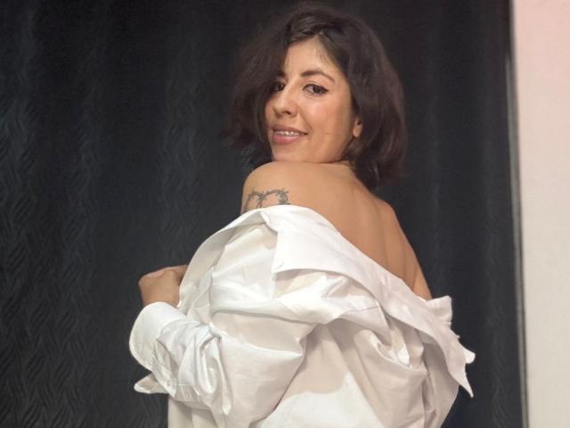 AnastasiaEvansX with short dark hair, smiling, white shirt off her shoulders, exposing her back and a small tattoo, standing against a dark patterned background.