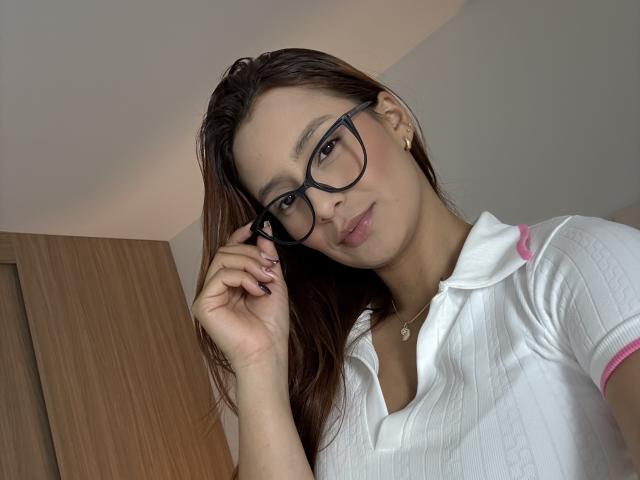 MyaSpecter with glasses, brown hair, white ribbed polo, pink trim, slightly parted lips, holding glasses, gold necklace, soft smile, light skin, subtle makeup, earring.