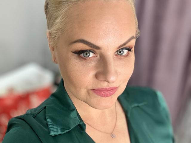 MermazingLight has blonde hair, green eyes, and is wearing a green satin shirt with pink lipstick. Her tits are slightly visible. She's looking at the camera with a smile.