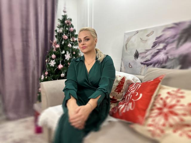 MermazingLight with blonde hair, green dress, sitting on couch, hands on knees, Christmas tree in background, purple curtains, red and white Christmas pillows, soft smile, relaxed.