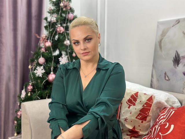 MermazingLight with blonde hair, green dress, crossed arms, sitting on couch, Christmas tree with pink ornaments in background, purple curtains, white wall, red and white pillow.