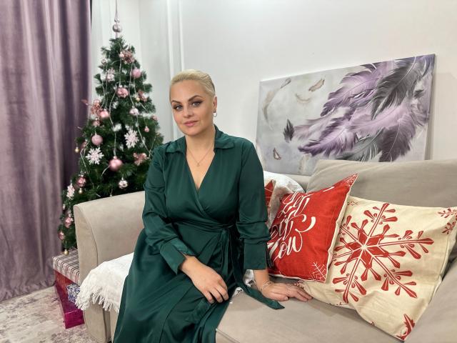 MermazingLight with blonde hair in a green dress, sitting on a beige sofa, has a cute ass, and is next to a decorated Christmas tree and red pillows.