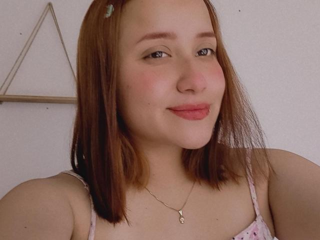 GabrielaMora with light brown hair, wearing a pink floral dress, has a cute smile, pink lips, small necklace, and a hair clip on the left side of her head.