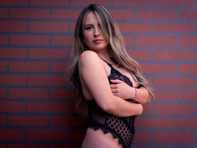 AmiiRosse with long blonde hair, wearing black lace lingerie, arms crossed, showing her tits, against a brick wall, looking at the camera with a slight smile.