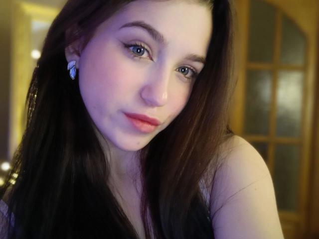 OliviaDesiree with long brown hair, blue eyes, and pink lips, wearing a black top, looking at the camera with a slight smile, wearing butterfly earrings.
