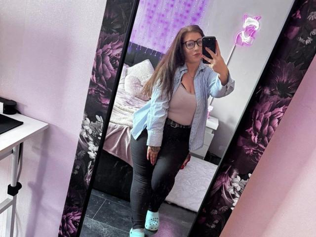 CurvyAlexaa – attraktiv mit Brille, langen braunen Haaren, einem hellblauen Hemd, einem pinken Oberteil, schwarzen Hosen und türkis gestreiften Socken, macht ein Selfie vor einem Spiegel und zeigt ihr Gesäß und ihre kleinen Brüste..