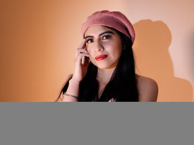 ZoeRedds has black hair, red lipstick, and a pink beret, touching her face with a smile, wearing a pink bralette, shadow on the wall, warm lighting.