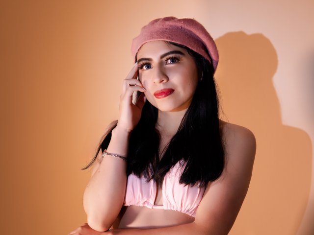 ZoeRedds has pale skin, black hair, and red lipstick, wearing a pink beret and matching bra, with a subtle smile, touching her cheek, shadow on a warm orange wall.
