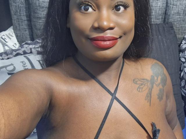 AlissaJordan with dark brown skin, red lipstick, and black hair, wearing a strappy top, showing a tattoo of wings on her right shoulder, and smiling at the camera.