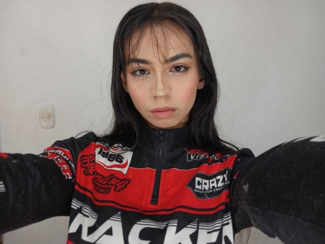 KatherinJohns with long black hair, wearing a red and black racing jacket, serious expression, reaching out with arms, visible eyeliner and subtle makeup, white wall in background.