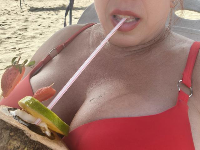 Khharha with big tits, red bikini top, biting pink straw from coconut drink with green, yellow, and red fruit, sandy beach background.