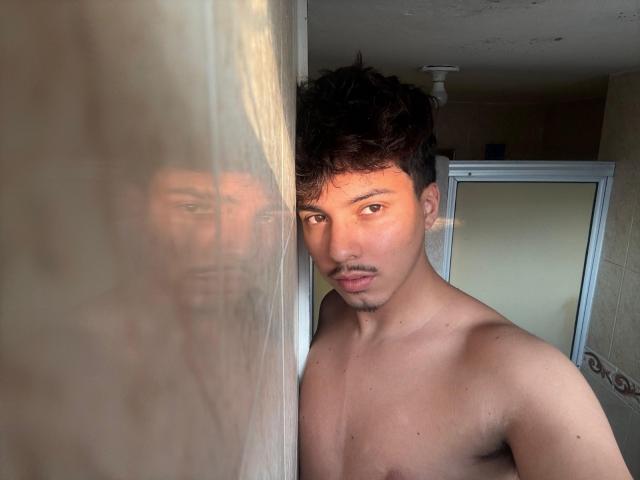 Shirtless SkySinclair with short brown curly hair and light brown skin, standing by a window, reflecting his face. His small, brown nipples are visible. He looks serious.