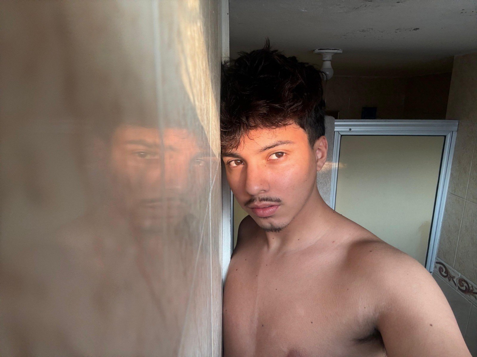 Shirtless SkySinclair with short brown curly hair and light brown skin, standing by a window, reflecting his face. His small, brown nipples are visible. He looks serious.