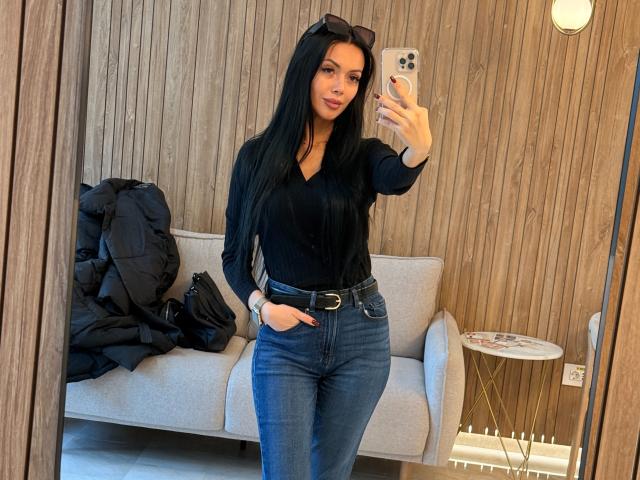 Nadeen with long black hair, wearing a black V-neck top and high-waisted blue jeans, standing in a mirror selfie, hands in pockets, holding a phone, wooden wall background.