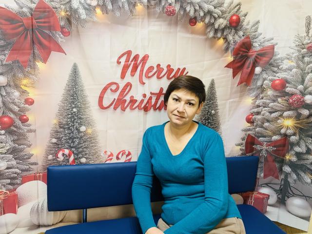 ZorynaYasna with short brown hair, blue sweater, and beige pants, sitting on blue bench, Christmas tree and ornaments in background, looking slightly to the side, small smile.