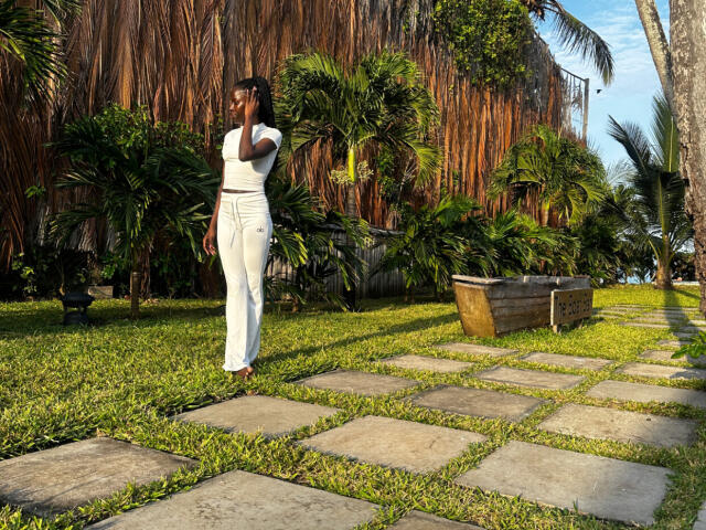 KharlaQueen stands in a sunlit garden, wearing a white, form-fitting dress that contrasts with her dark skin. She holds her right hand to her head, looking to the left. The garden has green grass, stone path tiles, and dense, tropical foliage. Tall palm trees and brown, hanging plant leaves form the background. A wooden bench is on the right. The image has warm, natural light.