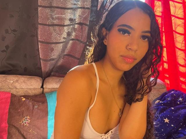 CherryCandyHot with curly brown hair, wearing a white tank top, showing her small tits, looking at the camera with a slight smile, in a warmly lit room.