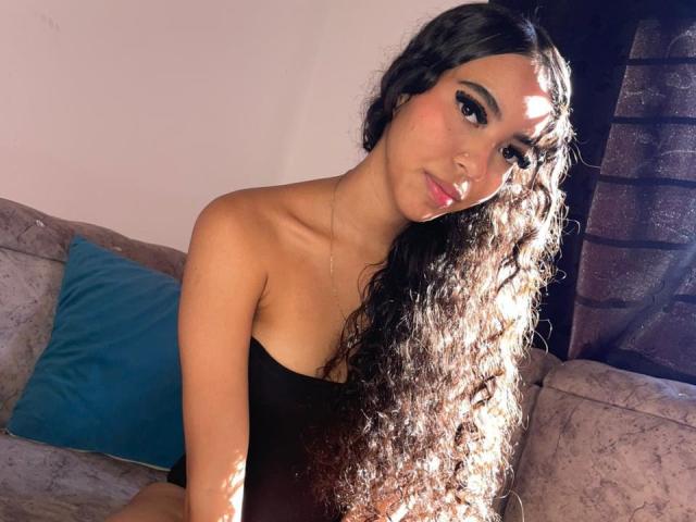 CherryCandyHot with long curly black hair, wearing a black strapless top, sitting on a couch with a blue pillow, sunlight highlighting her face and chest.