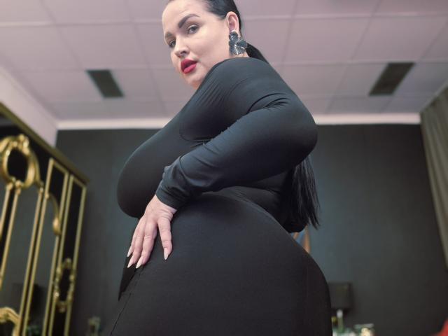 GeorgiaLerox with large, curvy ass in tight black dress, long black hair, red lipstick, and black earrings, looking over shoulder, hand on butt, ceiling tiles in background.