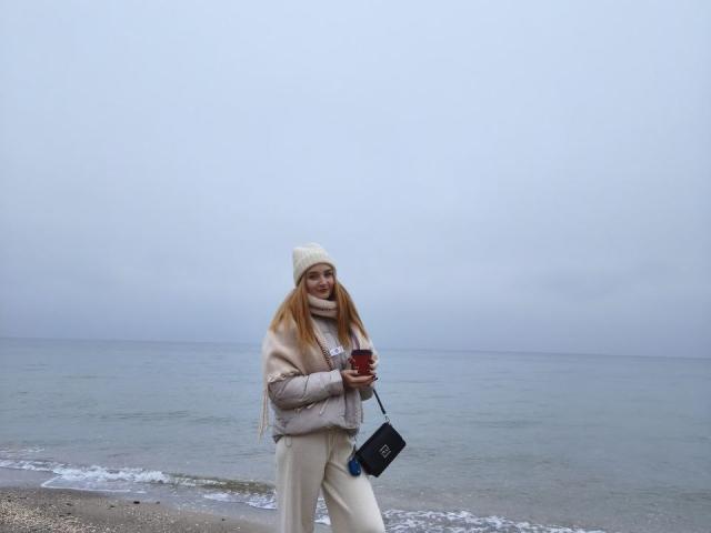 Celestiya, con cabello rubio largo, vestida con una chaqueta beige abultada, un gorro blanco y pantalones blancos, sosteniendo un bolso negro, de pie en una playa pedregosa con un océano brumoso..