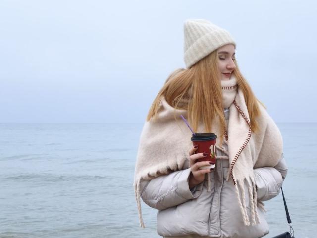 Celestiya, con una taza roja en la mano, una chaqueta blanca abultada, una bufanda beige y un gorro blanco, está de pie junto al océano, sosteniendo una pajita, mirando hacia un lado, y lleva una pequeña bolsa..
