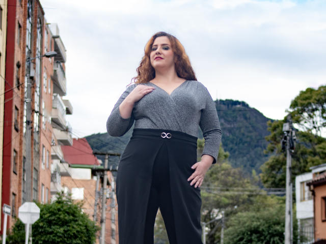 FreyaSeid, com cabelo ruivo cacheado, blusa cinza de decote em V, calça preta de cintura alta, em pé com confiança, com a mão no peito, mostrando os seios, em um cenário de rua urbana..