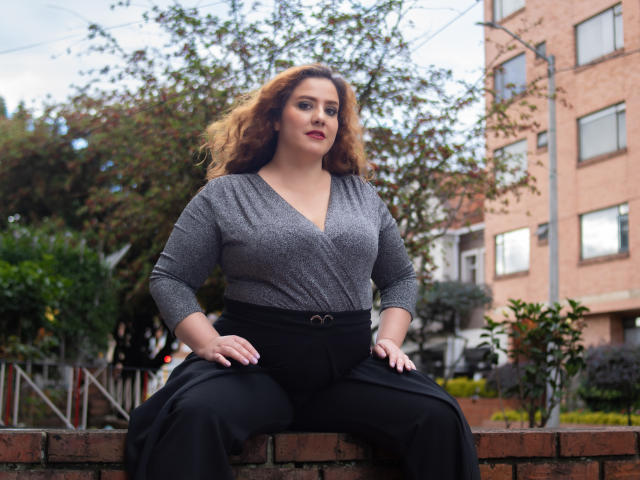 Freya, con cabello rojo ondulado, una blusa gris de cuello en V y pantalones negros, sentada en un muro de ladrillo, con las manos sobre los muslos, luciendo segura de sí misma. Se ven los senos, ligeramente grandes, rostro lindo, lápiz labial rojo..