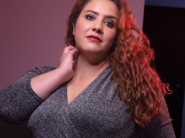 FreyaSeid with wavy red hair, wearing a gray textured top, has red lipstick and is touching her neck, looking slightly to the side. Boobs are medium-sized.