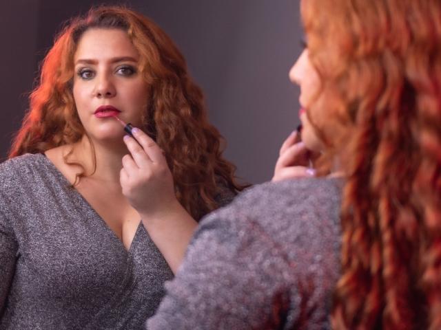FreyaSeid with curly red hair, gray dress, applying pink lipstick in mirror, showing her tits in reflection, holding lipstick in right hand, looking serious.