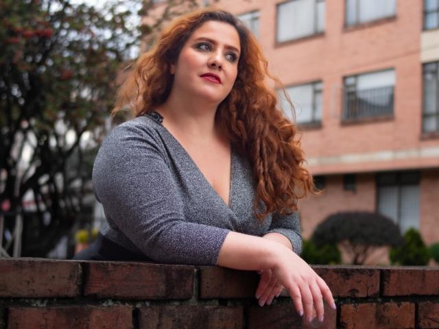 FreyaSeid has curly red hair, grey V-neck sweater, and red lipstick, leaning on a brick wall, looking up with a slight smile. Her ass is covered by the sweater.