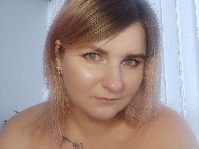MinistraX has pale skin, green eyes, and pinkish-brown hair, wearing small gold earrings, and a subtle tattoo on her neck, with a neutral expression and a slightly flushed face.