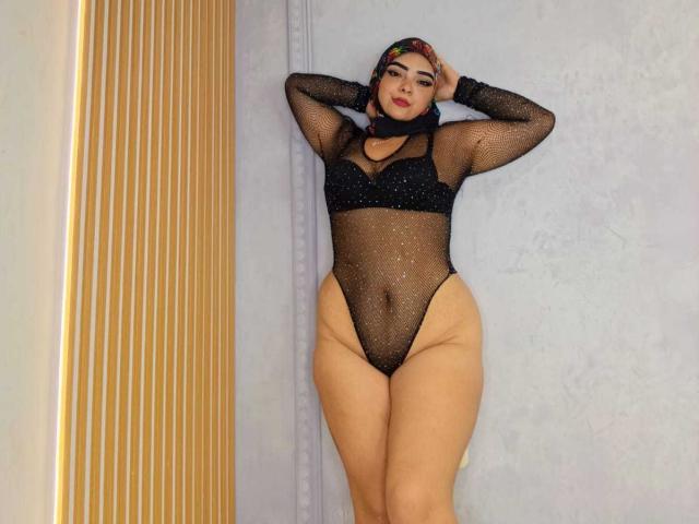 InayaKhalifa in a sheer black mesh bodysuit, showing her tits, pussy, and thick, curvy ass, with a colorful headscarf, hands behind her head, standing against a gray wall.