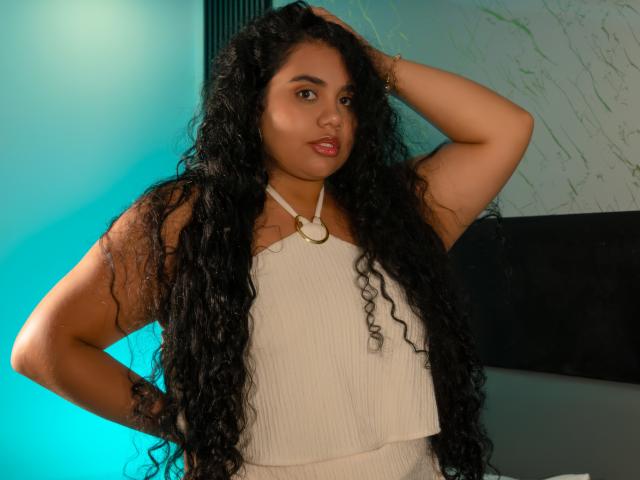 LisaTobon with long curly black hair, tan skin, and full red lips, wearing a white ribbed top with a gold ring, hand in hair, looking at camera.