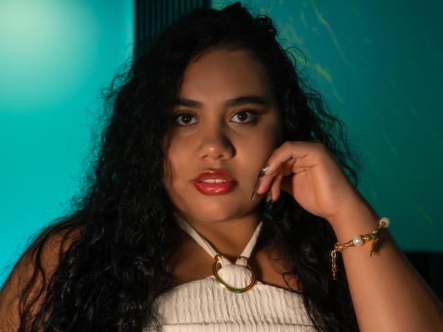 LisaTobon with curly black hair, red glossy lips, brown skin, wearing a white halter top with a gold ring, touching her cheek with a gold bracelet on her wrist.