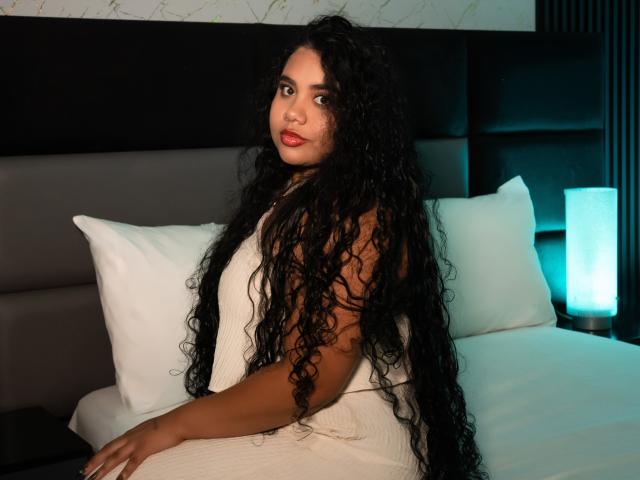 LisaTobon with long curly black hair, wearing a white dress, sitting on a bed with white pillows, looking over her shoulder, red lipstick, dark eyes, and a slight smile.
