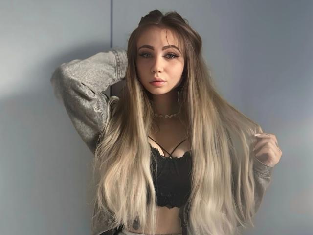 LilyLiebe-hot with long, blonde-to-brown ombre hair, wearing a gray jacket over a black lace bra, hand in hair, looking at camera, soft light, slightly parted lips.