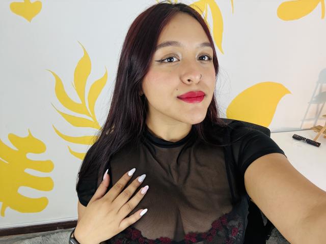 MarianaVargasX with dark red hair, black sheer top showing her tits, red lipstick, and white nails, holding her chest, yellow leaf wall art in the background.