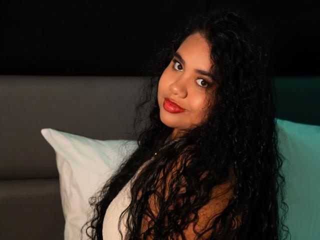 LisaTobon with long black curly hair, red lipstick, and brown eyes, wearing a white sleeveless top, sitting against a pillow, looking at the camera with a subtle smile.