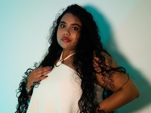 LisaTobon with long curly black hair, red lipstick, wearing a white sleeveless top, hand on chest, looking at camera, shadow on light blue wall, confident expression.