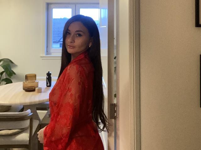 Dionisia with long brown hair, wearing a sheer red lace top, standing in a doorway, looking back, showing her tits, in a softly lit room.