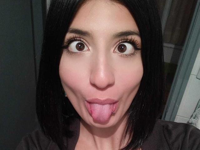 Kaiix has wide brown eyes, black hair, and is sticking out her pink tongue with a small nose piercing. Her long eyelashes are noticeable.