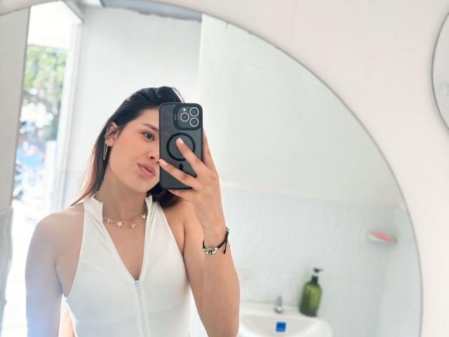 DannaHeat taking a selfie, pouty lips, white zip-up top, necklace, black phone, brown hair, in front of a mirror, holding phone, soft light, subtle makeup.