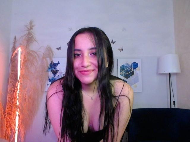 GabrielaMora with long black hair, pink lips, smiling, wearing a black top, showing small tits, necklace, and pink skin, leaning forward, standing in a softly lit room.