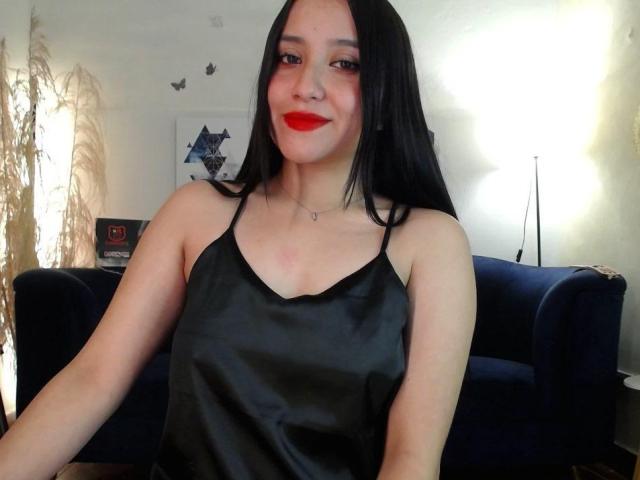 GabrielaMora with black hair, red lipstick, black satin camisole, showing her small tits, sitting in front of a blue velvet couch, smiling. She's wearing a delicate necklace.
