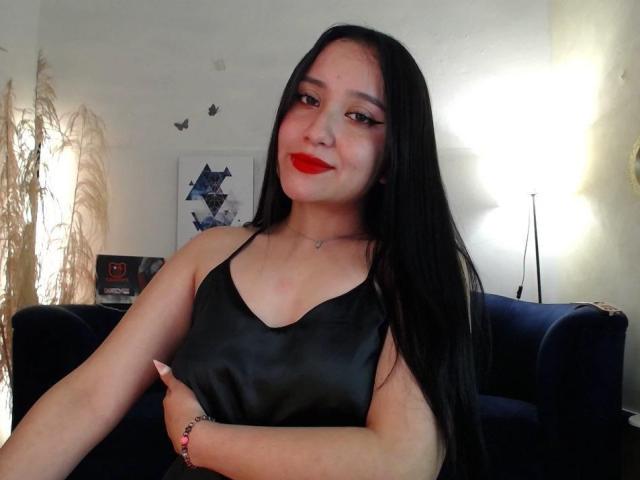 Gabriela Mora, con lunghi capelli neri, rossetto rosso, vestito di seta nera, che si tiene i seni, sorridendo, indossando un braccialetto, in piedi di fronte a un divano blu e una lampada..