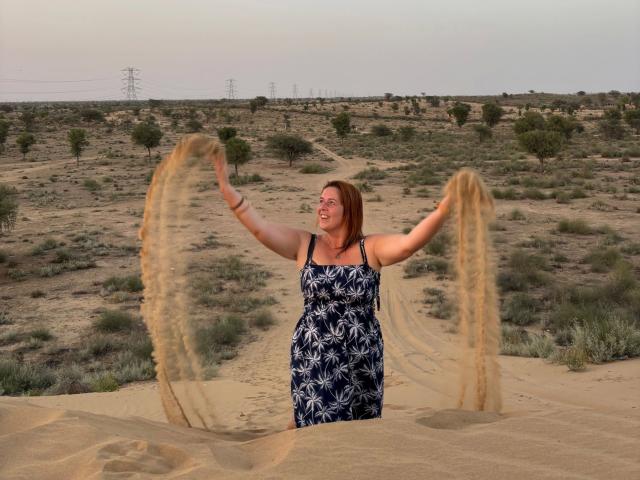 Chleo-hot with red hair, arms out, spinning a tan cloth in a sandy desert. She's wearing a blue floral dress, smiling, and has a cute, chubby ass.