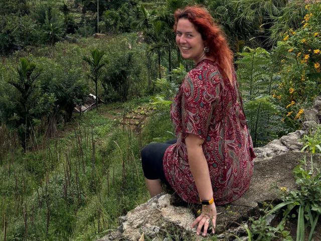 Chleo-hot with red hair, smiling, wearing a red patterned shirt and black pants, sitting on a rock, looking back over her shoulder, surrounded by green foliage.