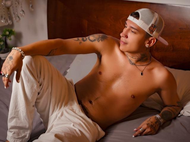 HappyEndd, ohne Hemd, weiße Hose, Mütze verkehrt herum, Tattoos, Halskette, Armband, Ring, liegt im Bett, Arm über dem Knie, blickt zur Seite, Bett mit grauen Laken, entspannt, mittelgroßer Körperbau, süß..