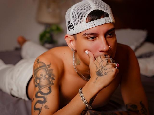 HappyEndd ohne Hemd, mit Tattoos, weißer Mütze, Nasenring und Kette, liegt im Bett, Hand in der Nähe des Mundes, weiße Hose, Armbänder und Tattoos am Arm und an der Hand, blickt in die Kamera..