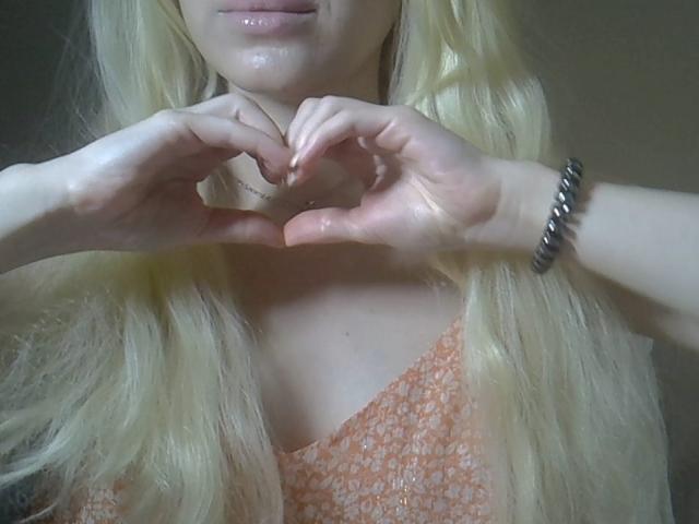 DitaVira with long blonde hair, hands making a heart shape, wearing an orange floral dress, and a silver bracelet, showing her glossy pink lips.